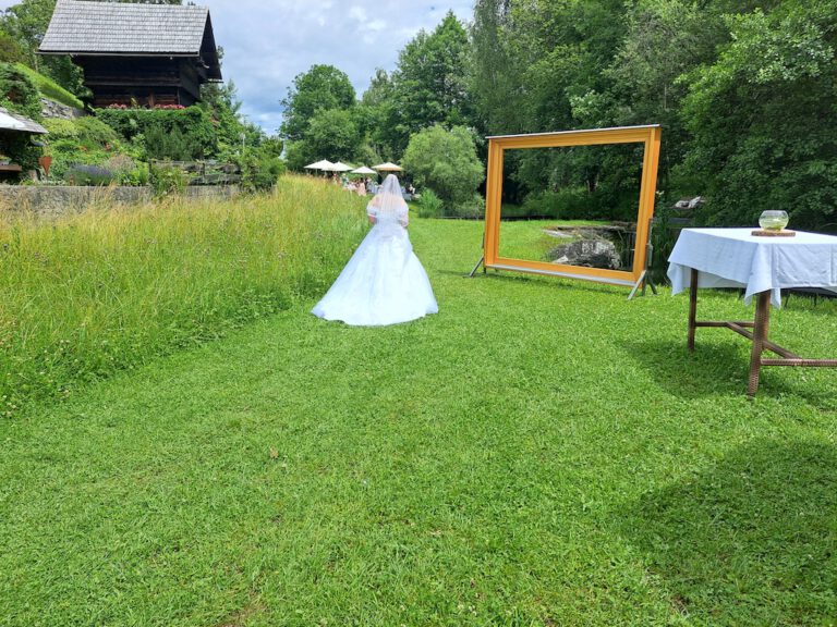 Braut in weißem Kleid schreitet über eine grüne Wiese zu einer Hochzeitszeremonie im Garten, mit dekorativem Holzrahmen und Naturkulisse.