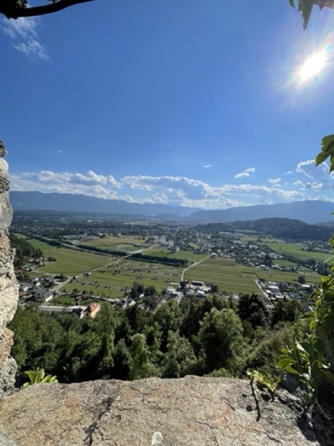 Aussicht über das Drautal mit Feldern, Ortschaften und umliegenden Bergen unter blauem Himmel bei Sonnenschein.